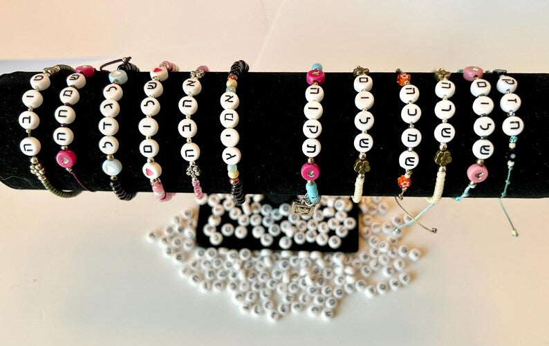 DIY Hebrew letter bracelet making kit with seed beads and silver charms by Pretty Beans, featuring colorful beads and jewelry supplies.
